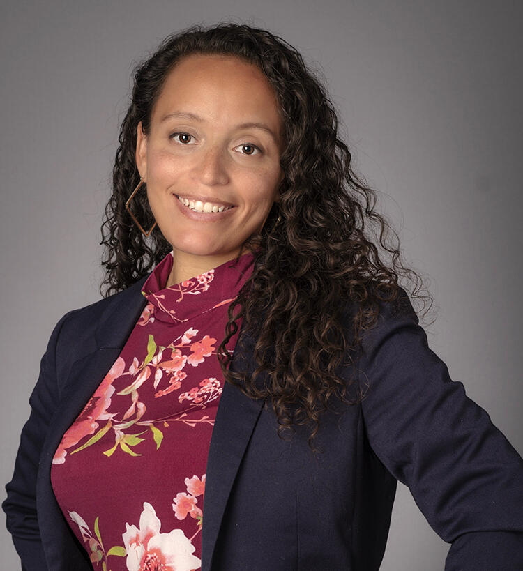 Dr. Hayley smiling looking directly at camera with long curly hair passing her shoulders, hoop earrings in a navy blazer with floral magenta top.